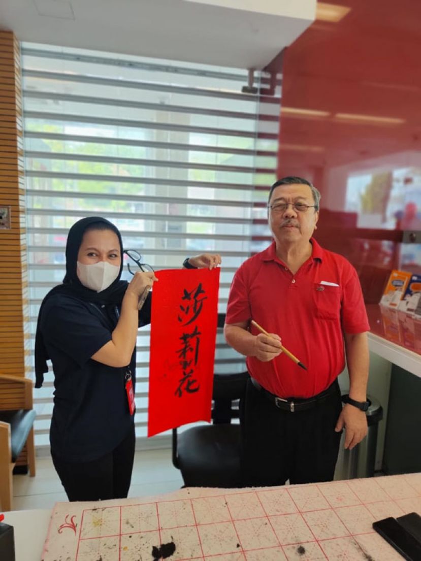 古晋书画研究会名誉会长许庆喜（右）在该活动上与丰隆银行槟岭分行友族职员展示挥毫作品。
