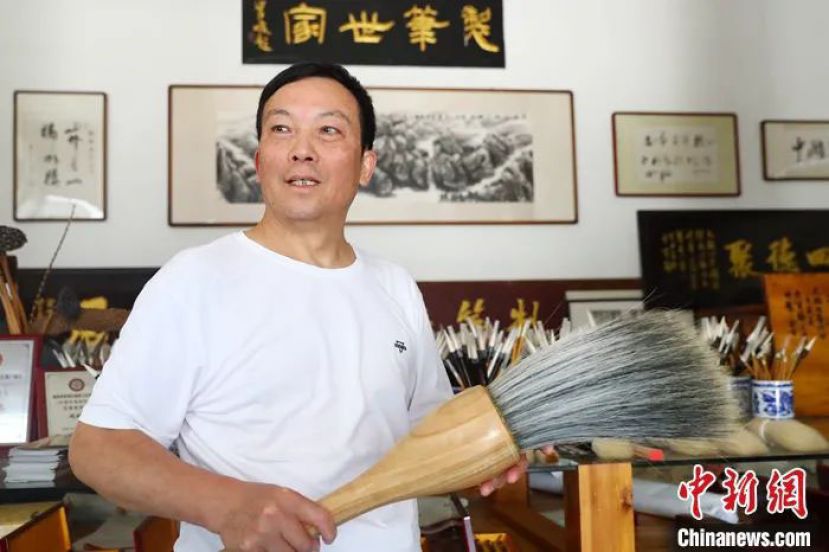 周鹏程手中拿着一支巨型毛笔，身后的牌匾上写着&ldquo;制笔世家&rdquo;四个字。